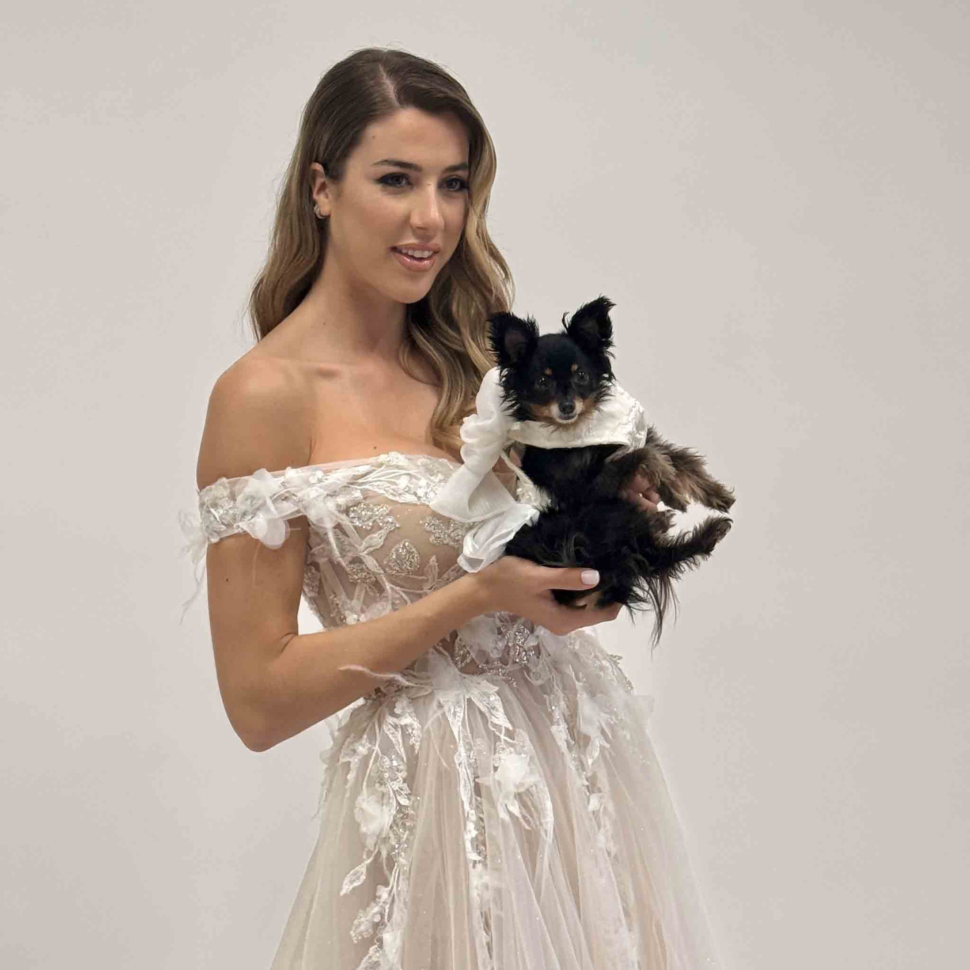 A woman in a white, off-the-shoulder lace dress is holding a small black dog. The dog is dressed in a white outfit that complements the woman's attire.